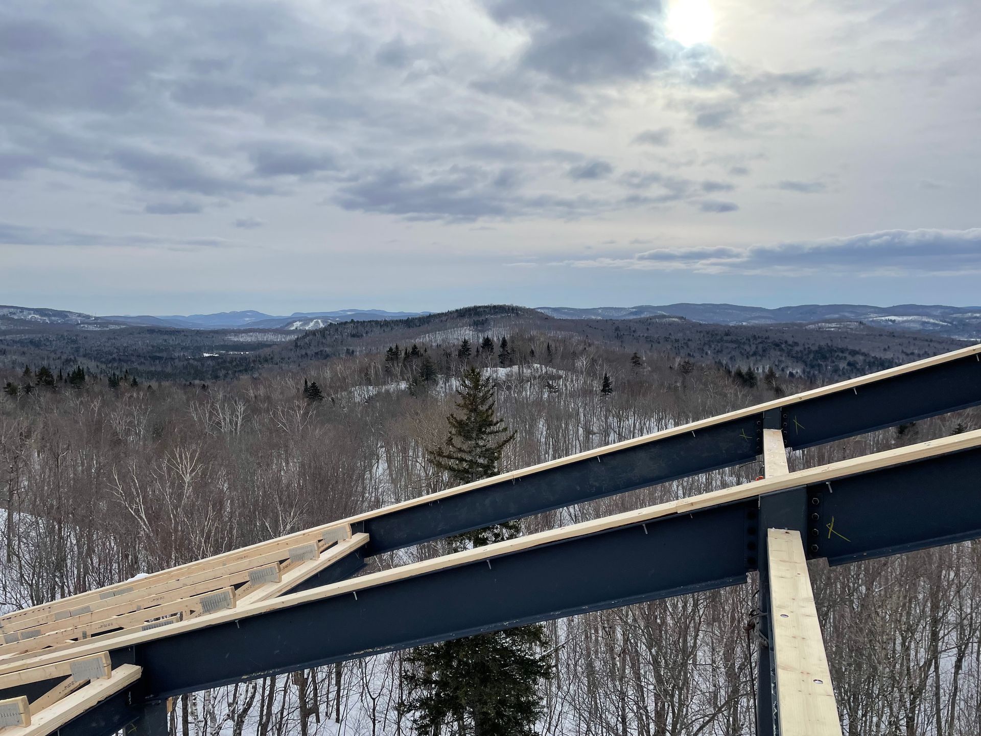Paysage enneigé vu à travers des poutres métalliques. Collines et arbres sous un ciel nuageux.