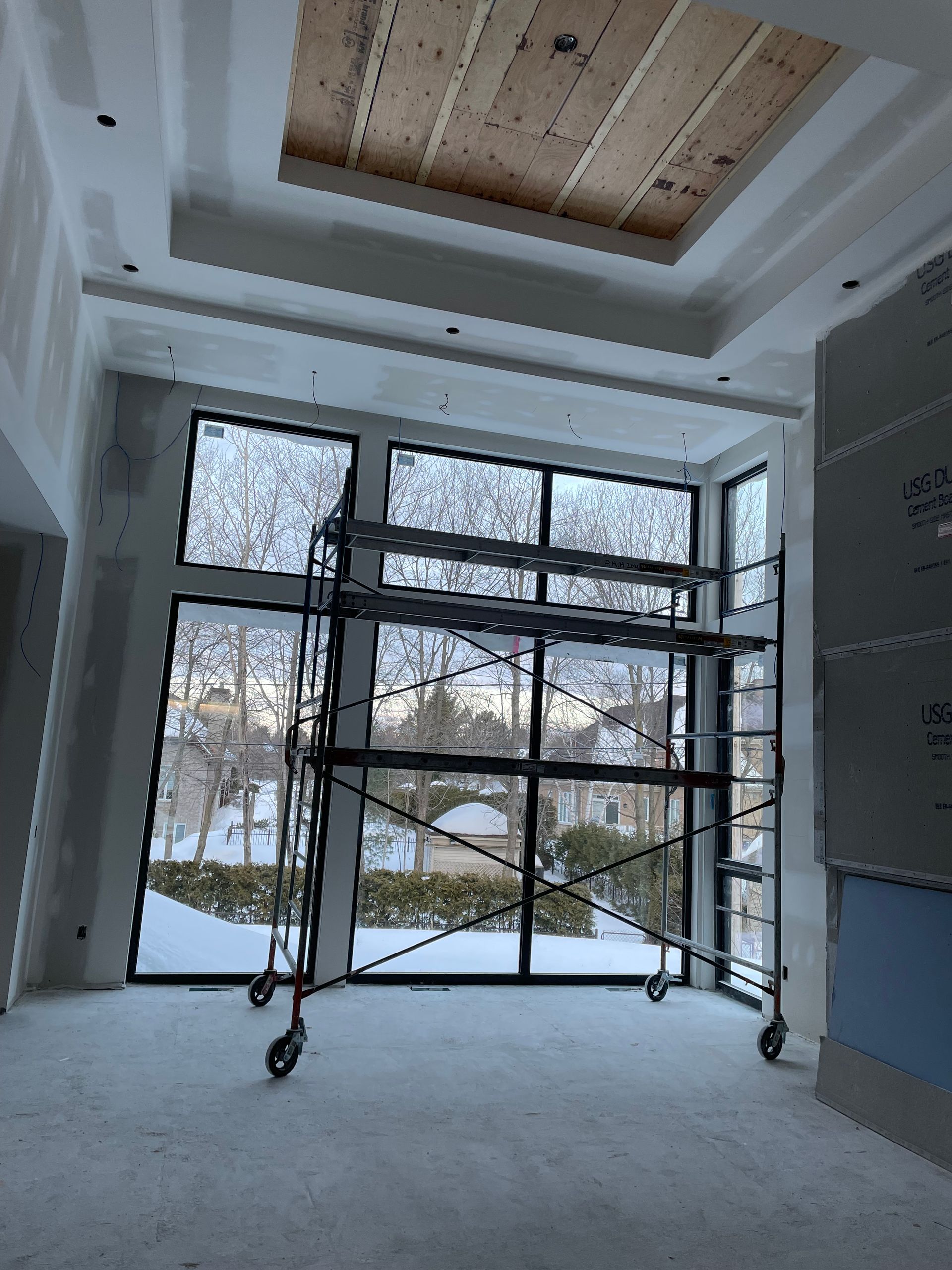 Intérieur en travaux avec des échafaudages près des grandes fenêtres, des arbres visibles et un plafond en retrait.