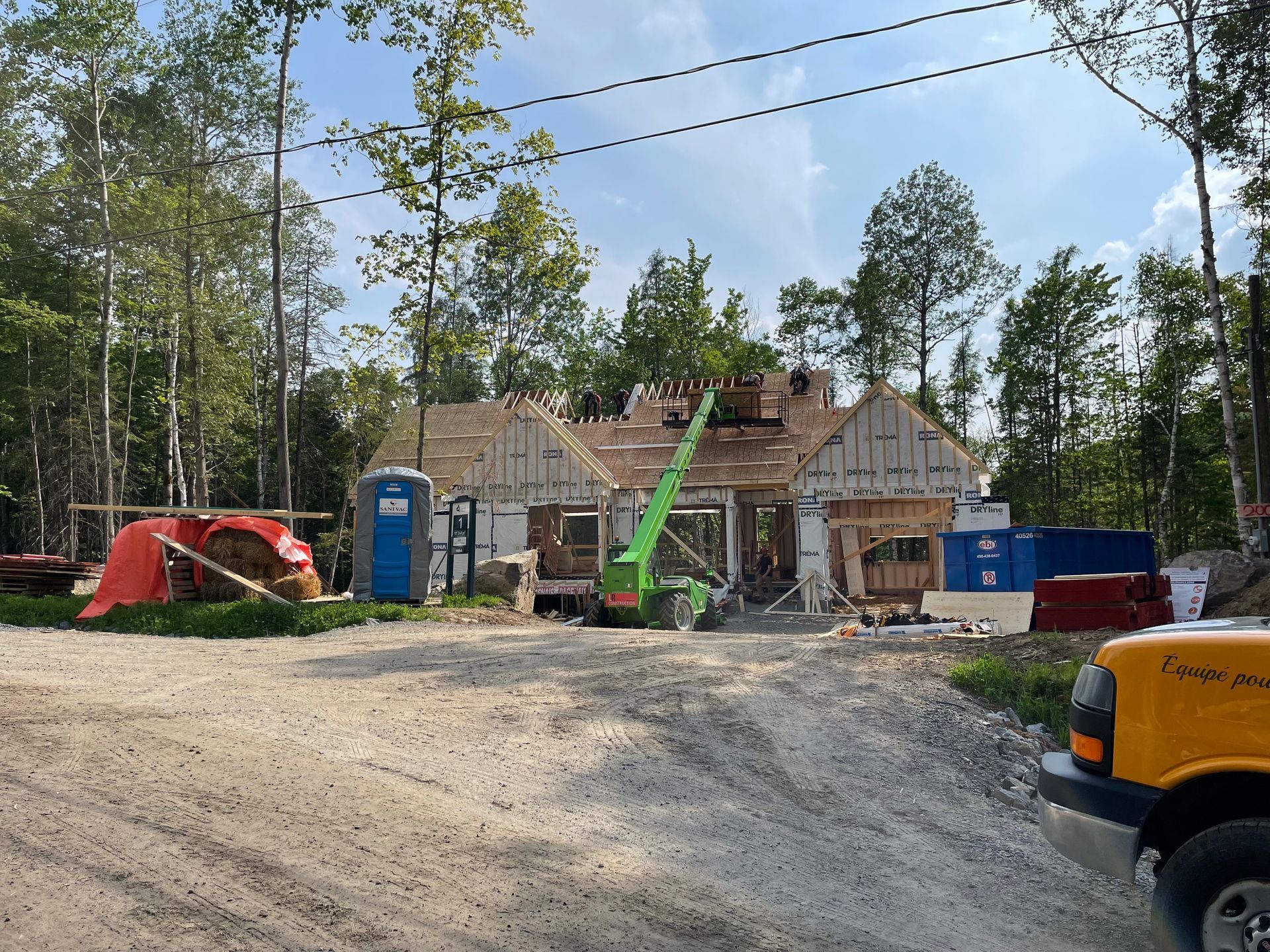 Chantier de construction avec une maison partiellement construite, des arbres en arrière-plan, des toilettes portables et une grue.