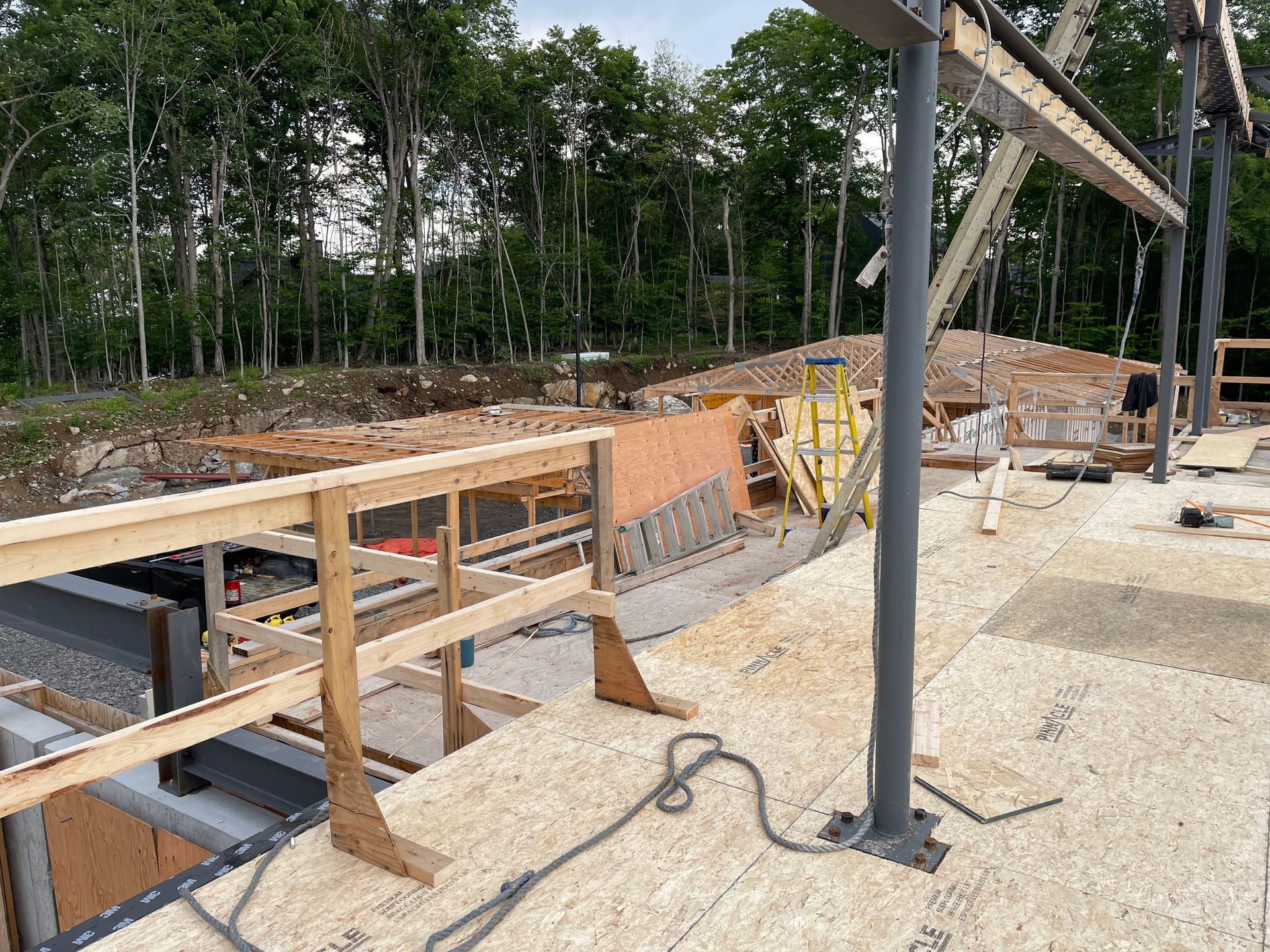 Chantier de construction : rambardes en bois sur une terrasse, poutres de soutien métalliques, charpente inachevée, arbres en arrière-plan.