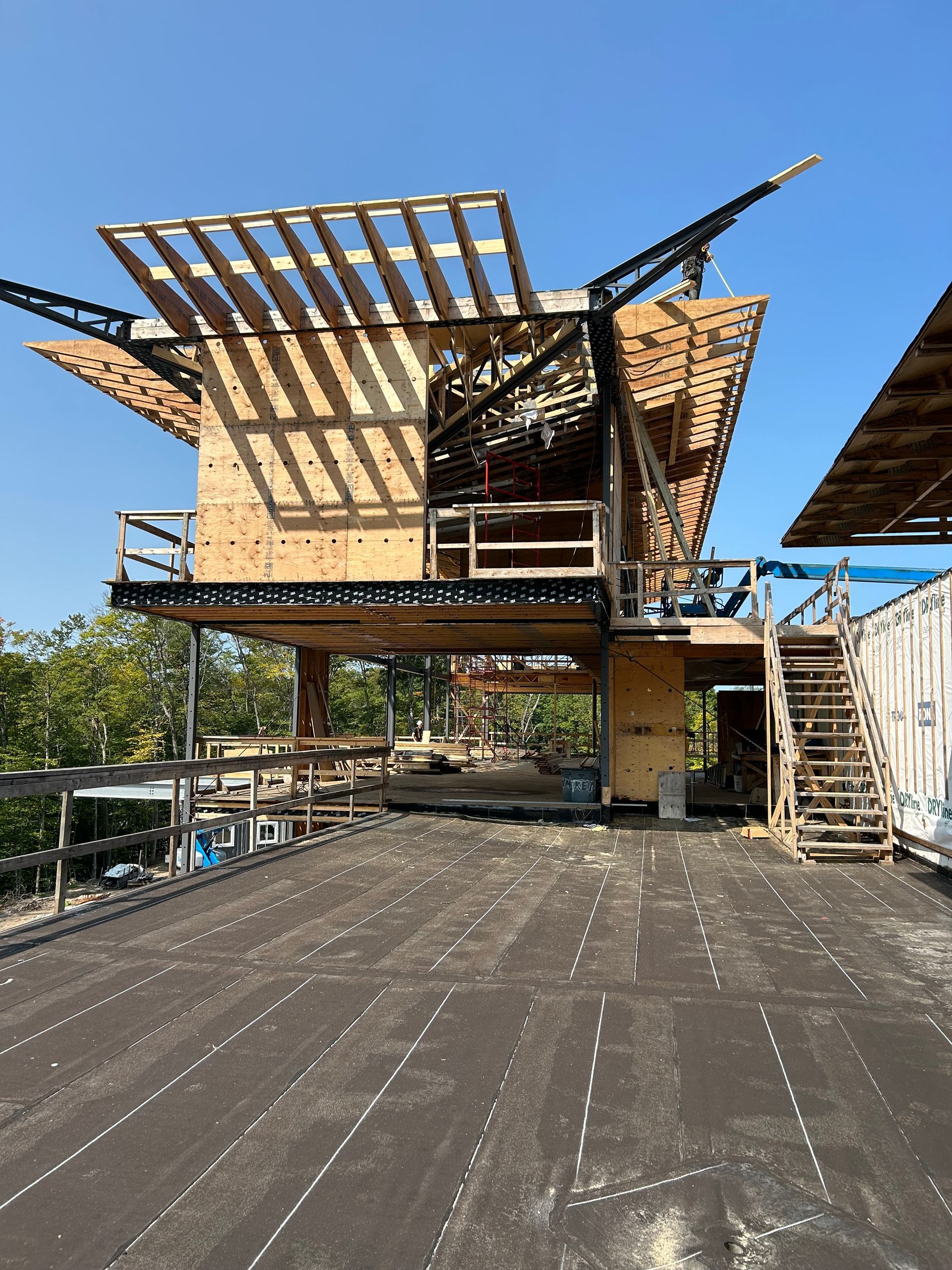 Un bâtiment moderne en bois en construction, avec un toit en surplomb et des escaliers, situé dans un environnement extérieur ensoleillé.