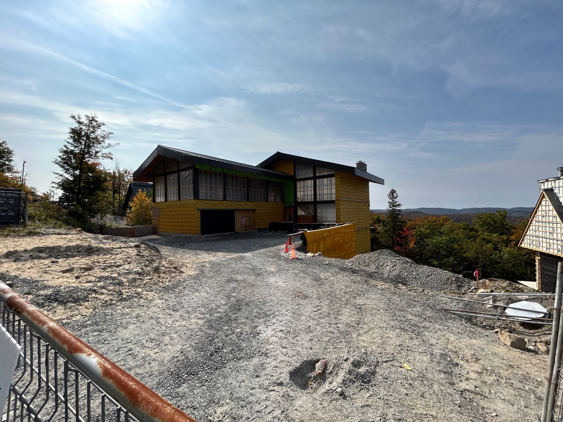Maison jaune en construction, allée de gravier et arbres environnants sous un ciel bleu.