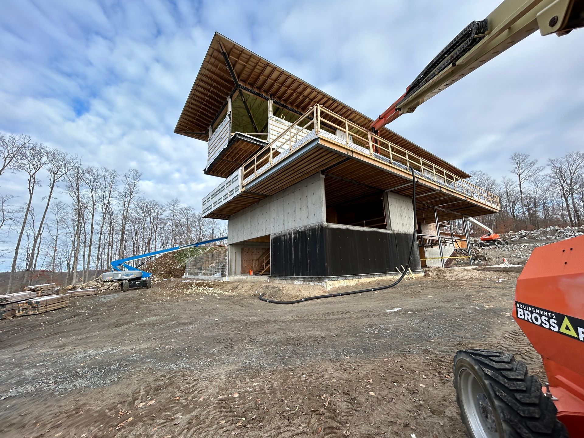 Bâtiment en construction, à ossature bois et base en béton, entouré d'arbres et d'engins de chantier.