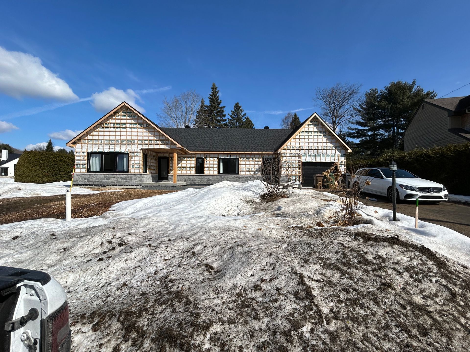 Maison en construction avec charpente apparente, cour enneigée, ciel bleu azur.