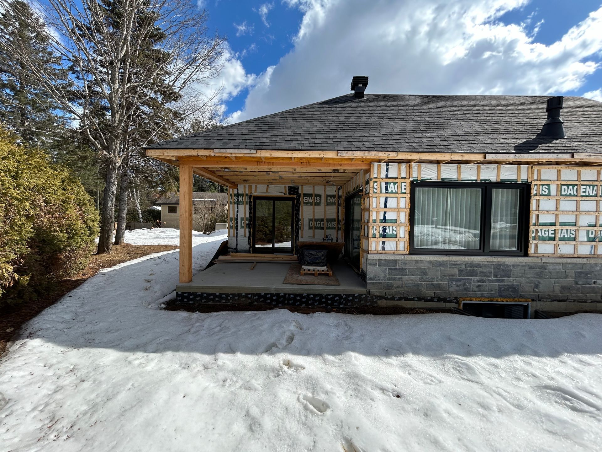 Maison en construction avec des murs apparents, de la neige au sol et un porche partiellement construit.