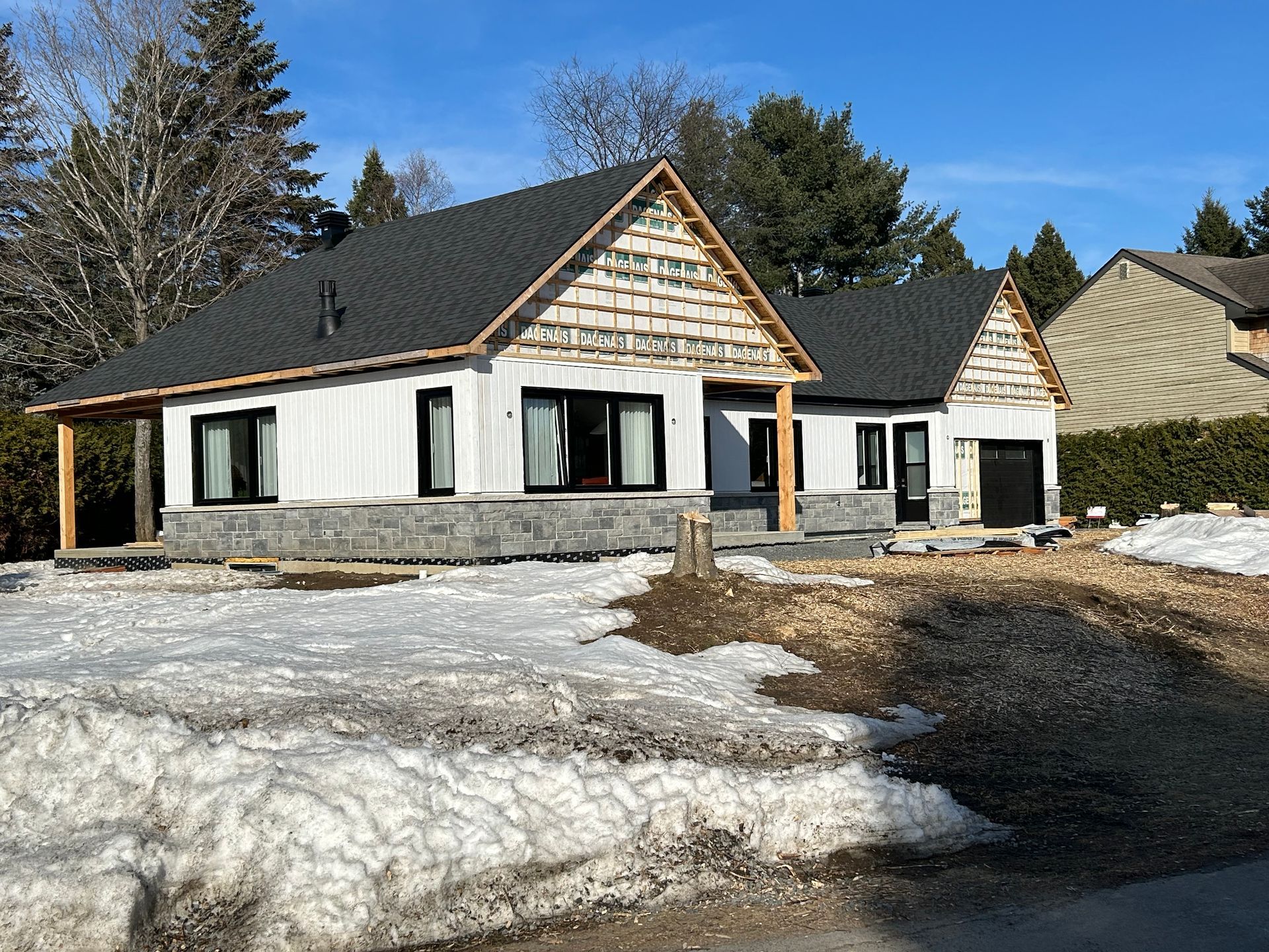 Maison en construction avec toit noir, poutres en bois, bardage blanc et fondations en pierre ; neige au sol.