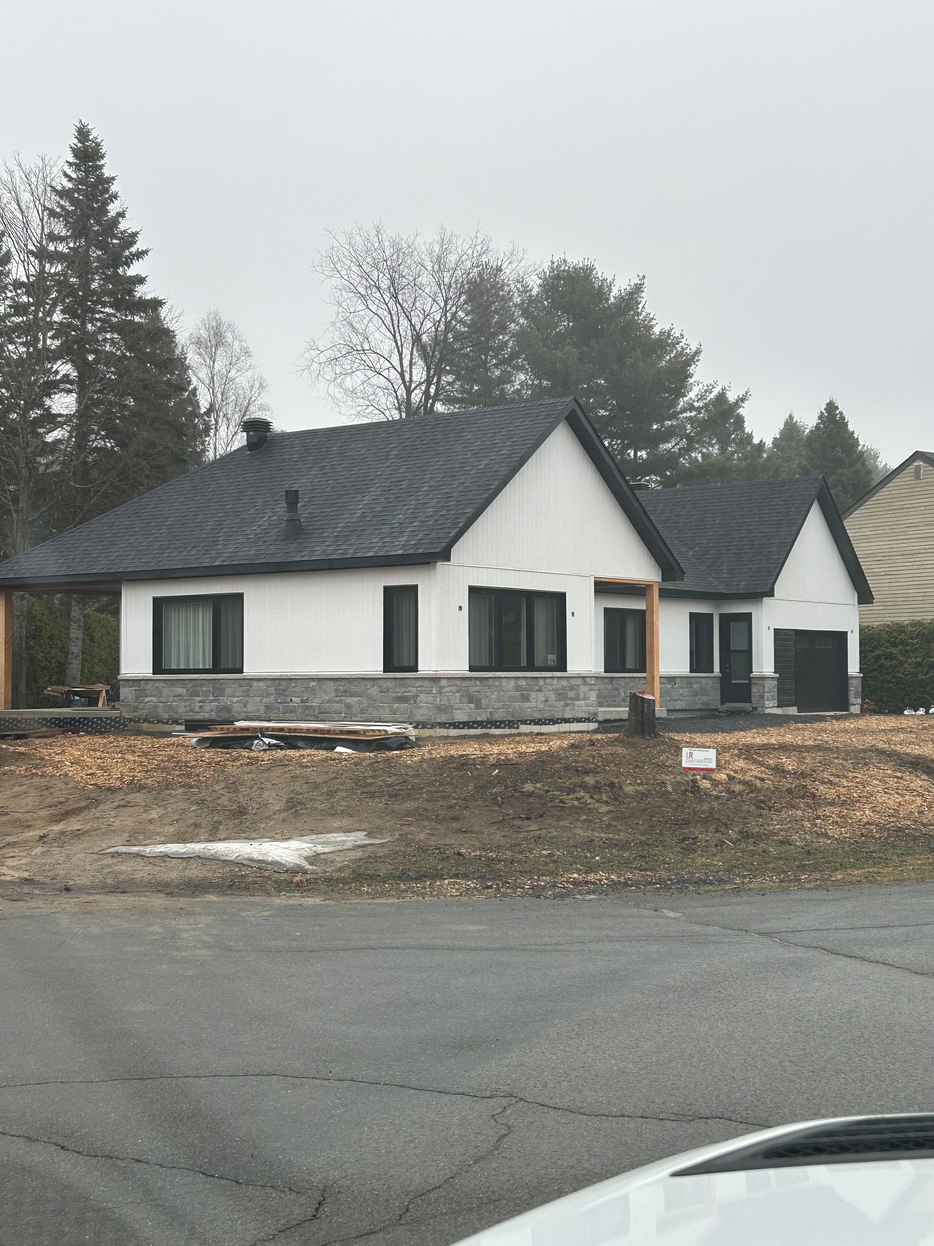 Un bungalow blanc au toit gris foncé se dresse par temps nuageux. Façade en pierre à sa base. Arbres en arrière-plan.