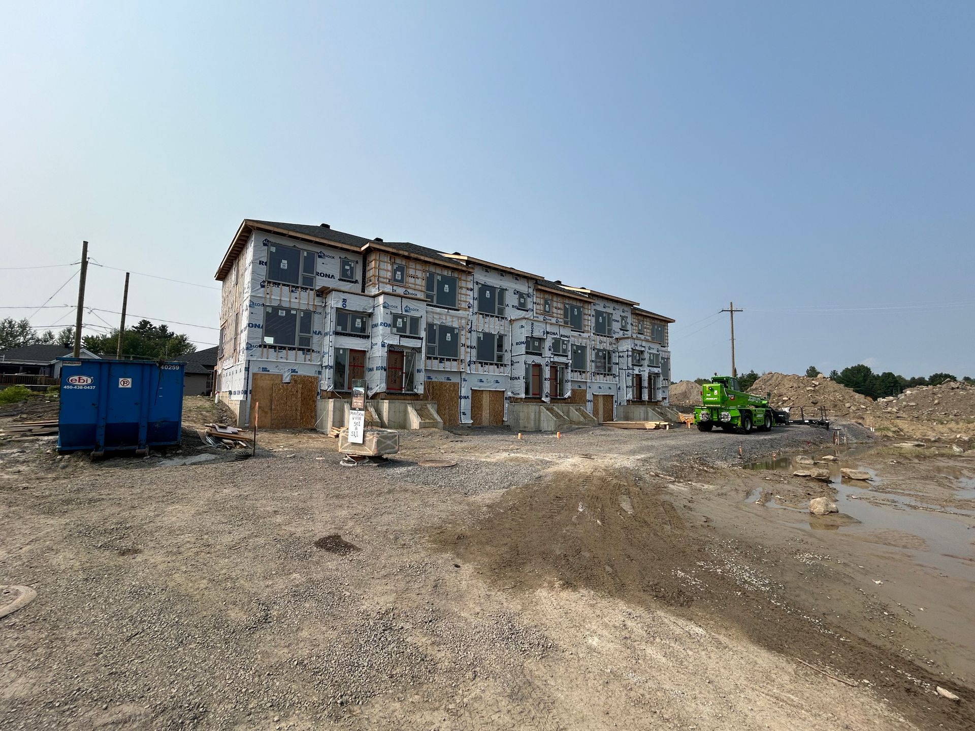 Maisons de ville en construction, recouvertes d'un pare-vapeur, sur un terrain de gravier sous un ciel bleu.