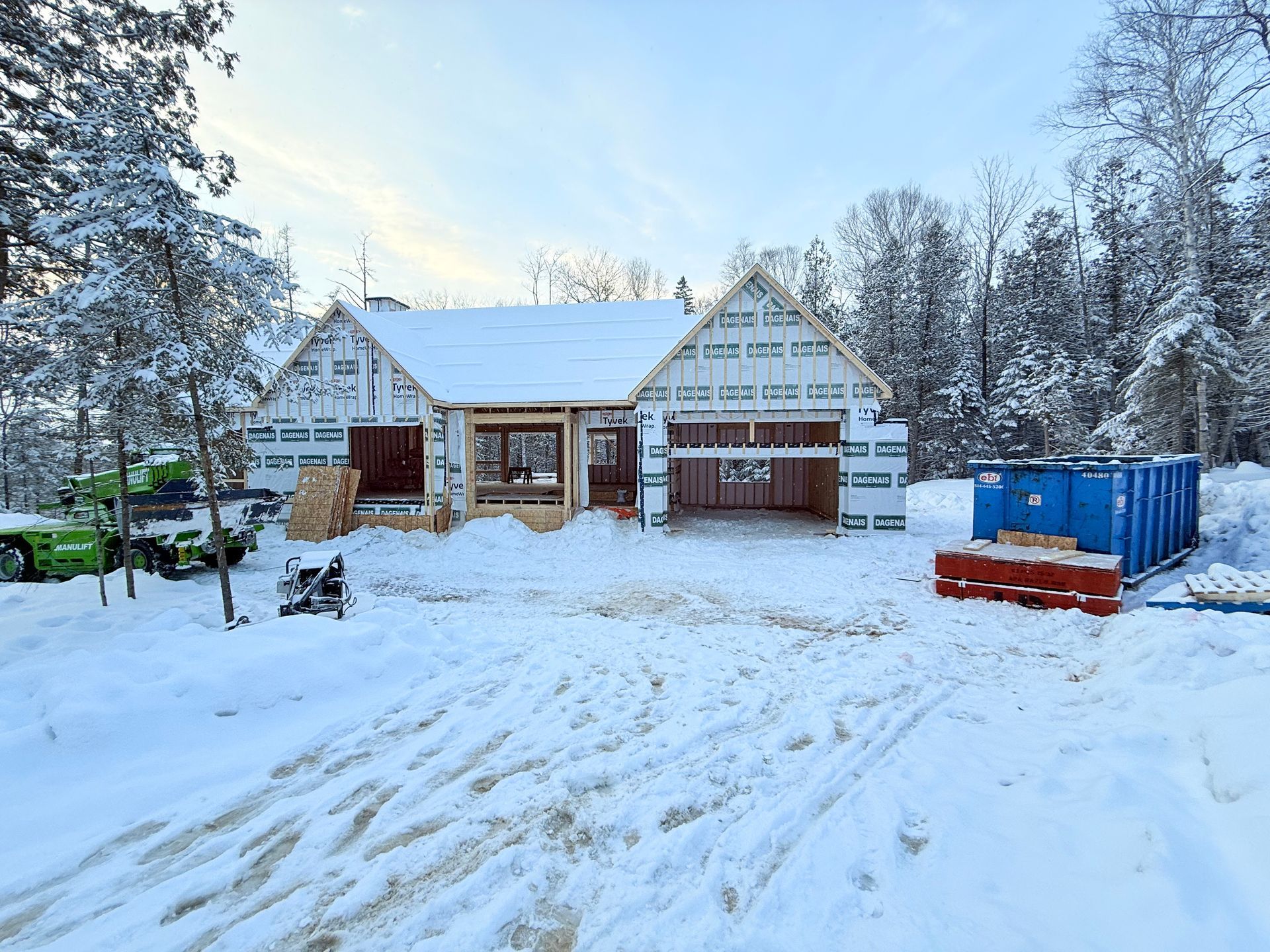 Maison en construction dans un paysage enneigé ; charpente en bois visible, benne à ordures bleue et engins de chantier verts.