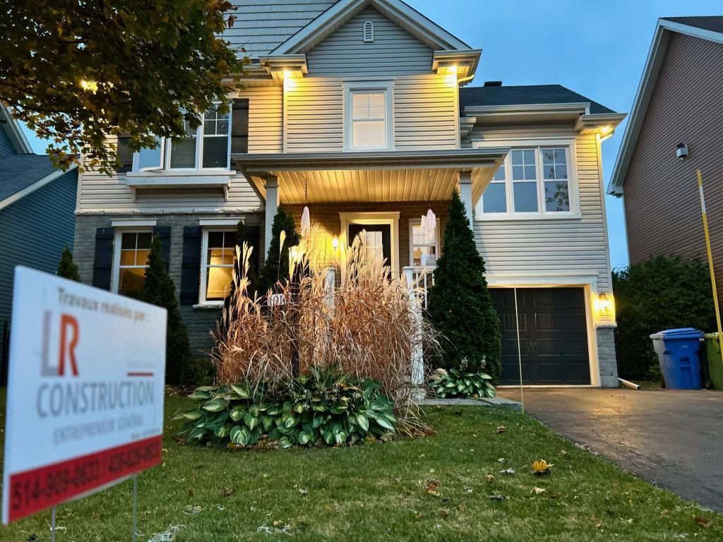 Maison à deux étages avec bardage gris, porte d'entrée éclairée, panneau LR Construction sur la pelouse.