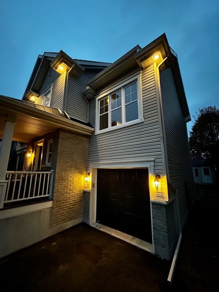 Maison à deux étages avec éclairage extérieur ; porte de garage fermée. Bardage gris, accents de brique, ciel bleu.