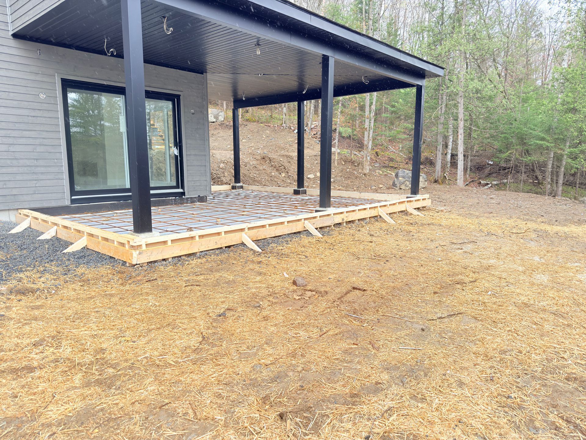 Terrasse en bois avec toit de porche, piliers noirs, surface en gravier et cour gazonnée.