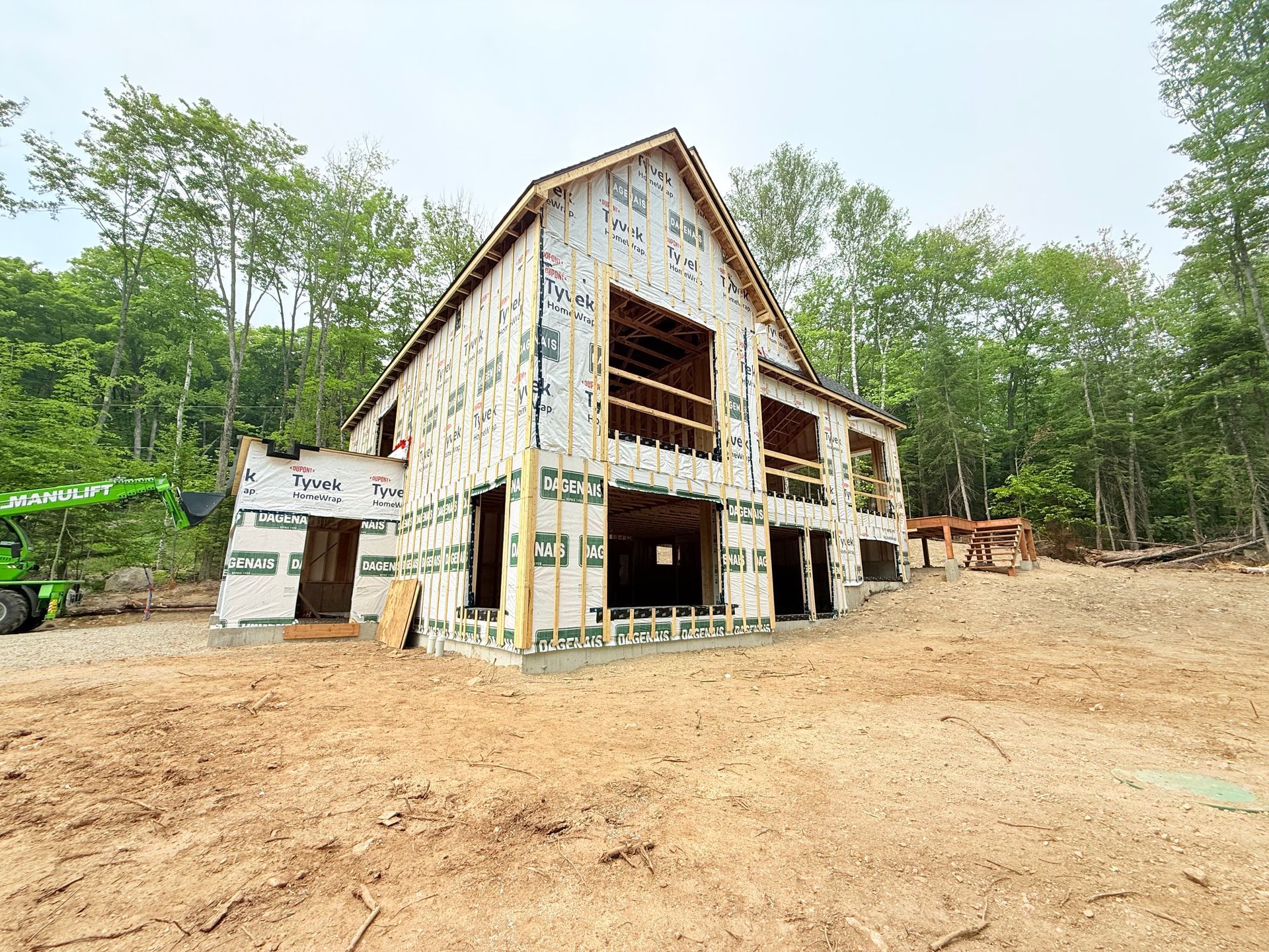 Maison à deux étages en construction dans une zone boisée, à ossature bois apparente et recouverte de matériaux de construction.