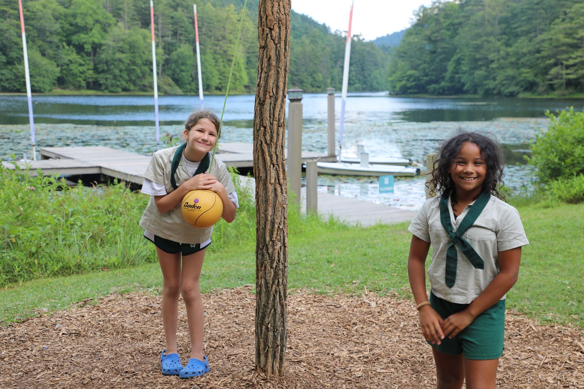 Two people play tetherball on a grassy field near a lake with kayaks on a dock in the background.