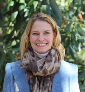 A smiling person with blonde hair wears a light blue blazer, grey turtleneck, and a brown leopard-print scarf outdoors.