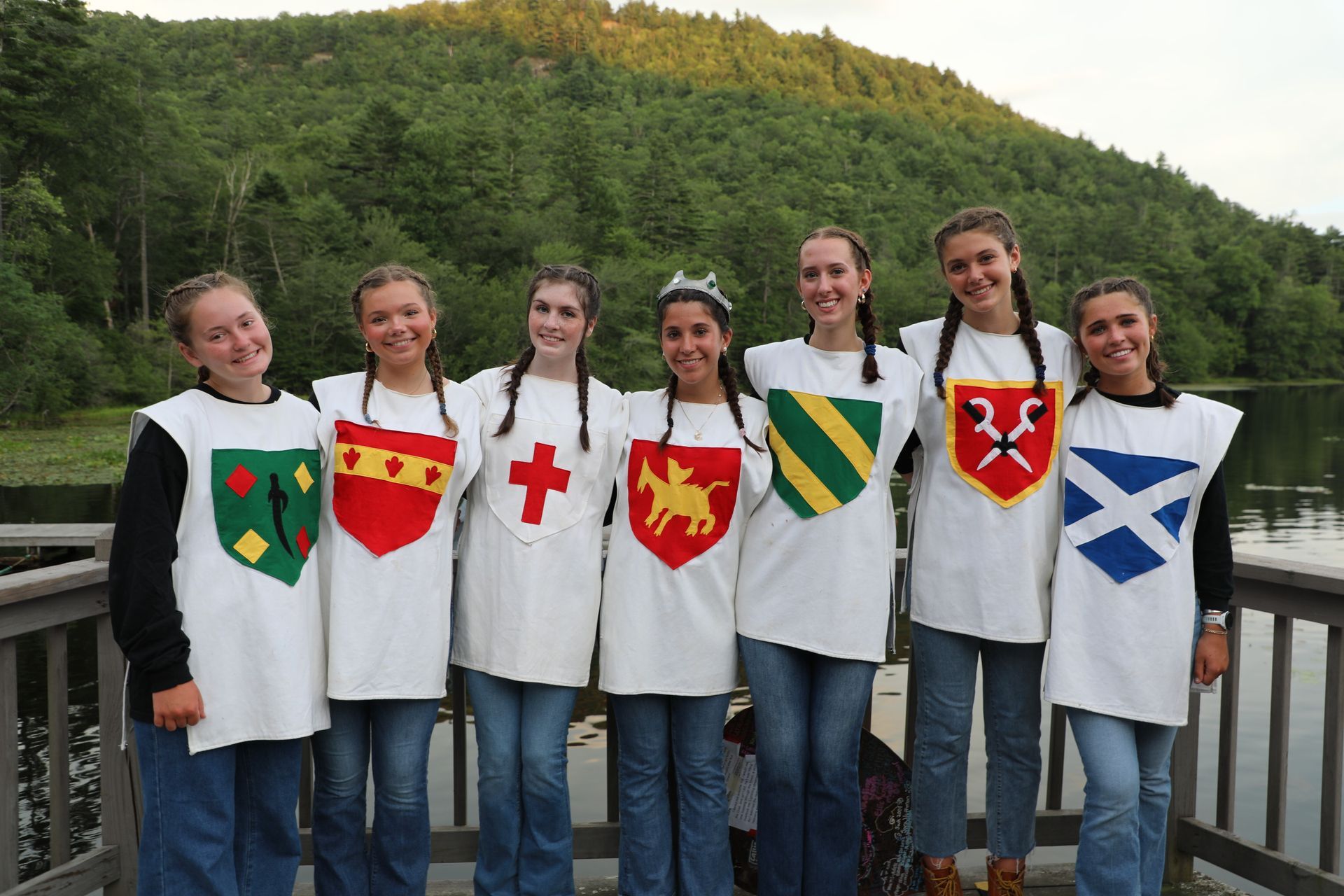 Seven people standing on a dock wearing white tabards with unique heraldic shields, backed by a lake and green hillside.