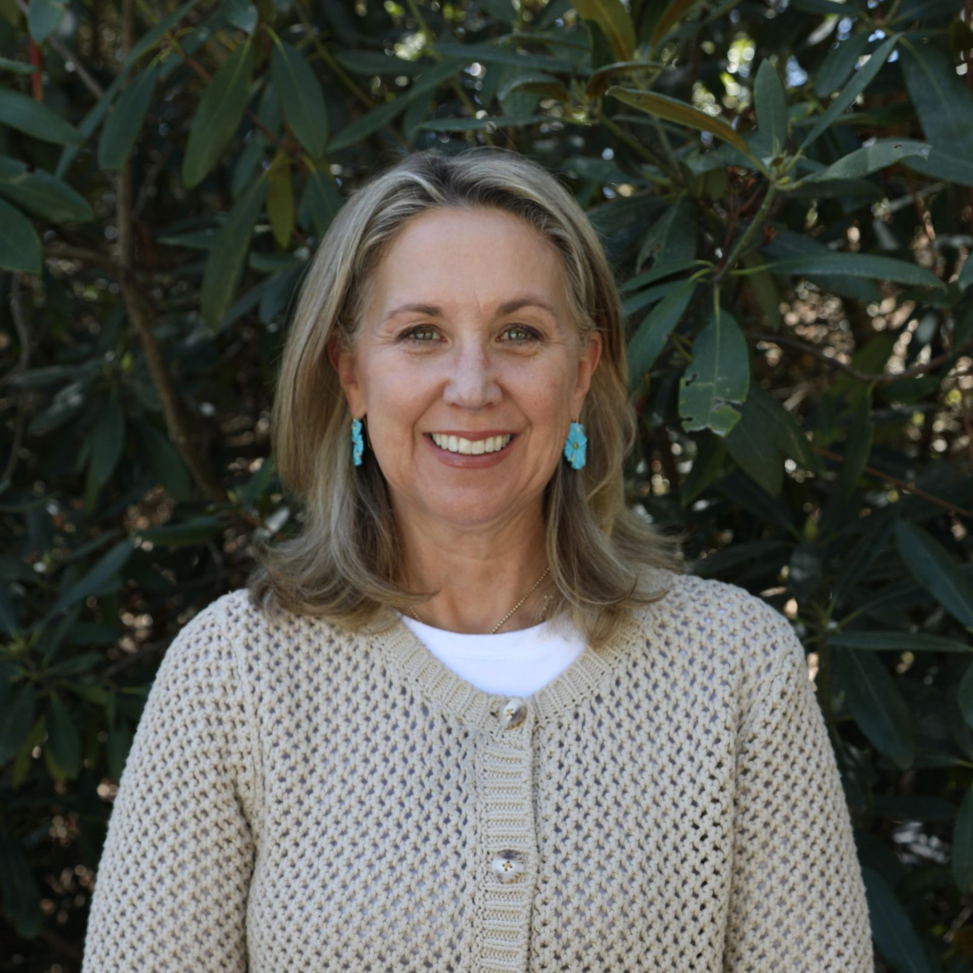 A smiling woman with blonde hair, wearing a dark maroon quilted jacket, standing in front of a stone wall and tree.