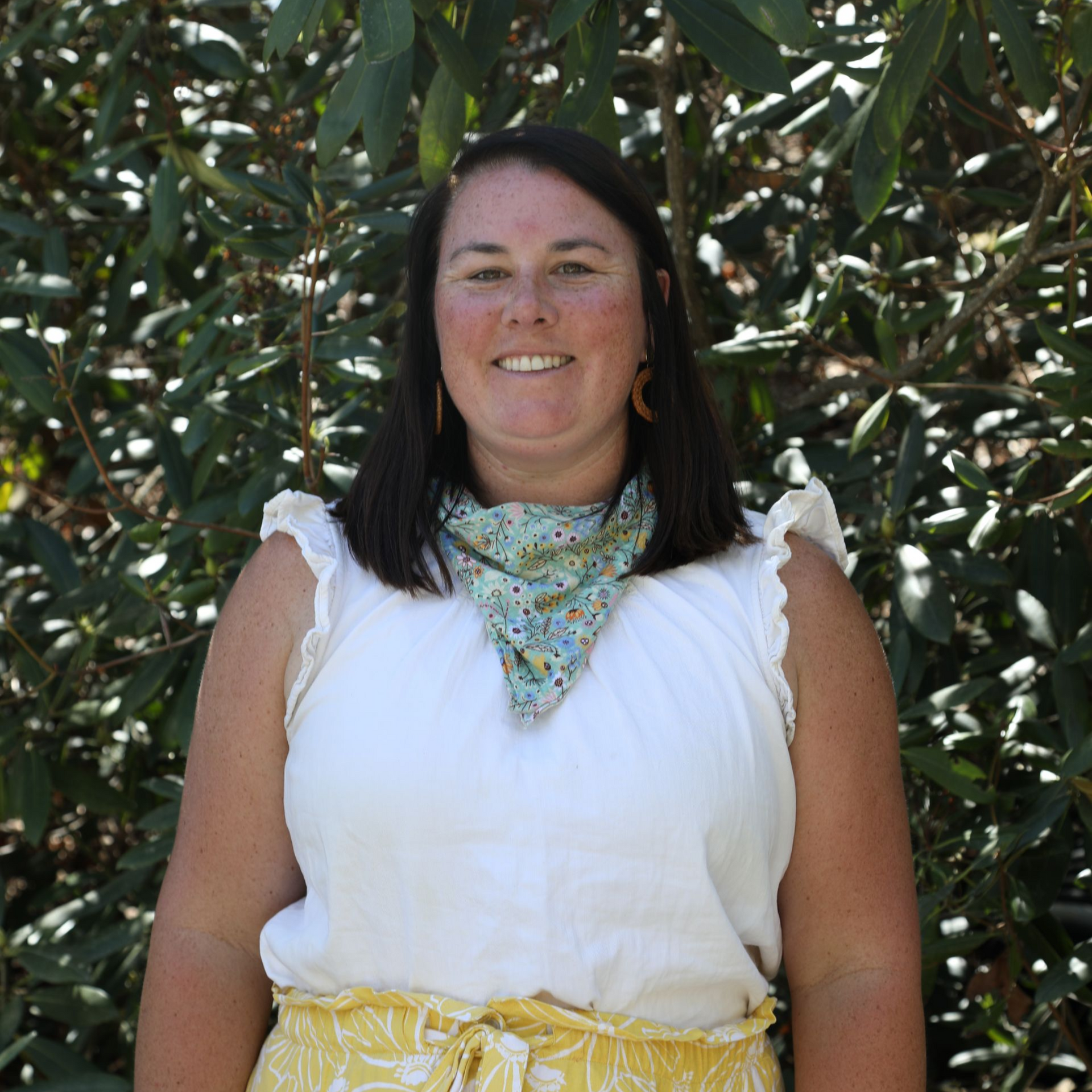 A person smiles while wearing a patterned neck scarf, a black shirt, and a light blue open button-down shirt outdoors.