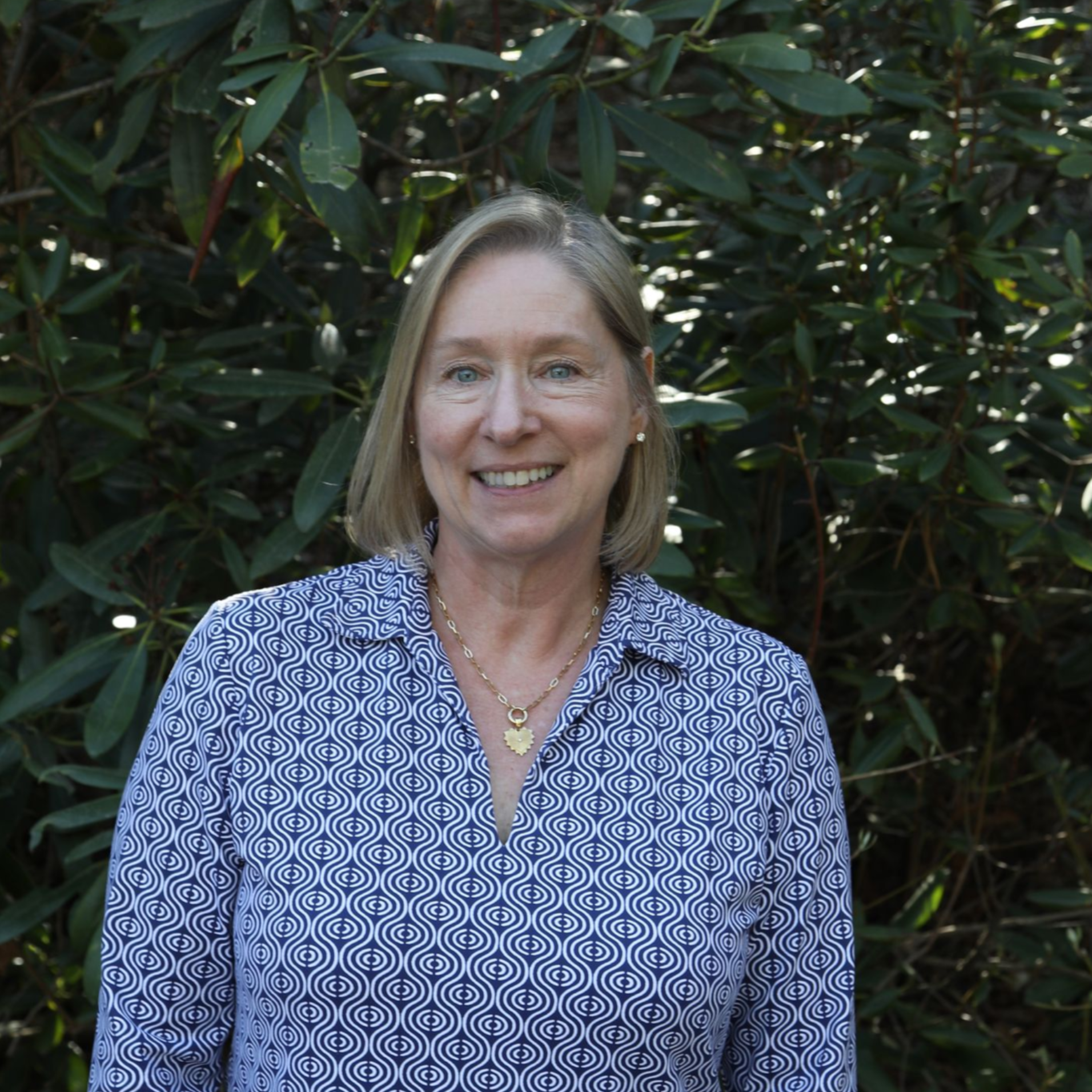 A woman with shoulder-length blonde hair, wearing a black blazer and a pearl necklace, smiling against a blurred background.