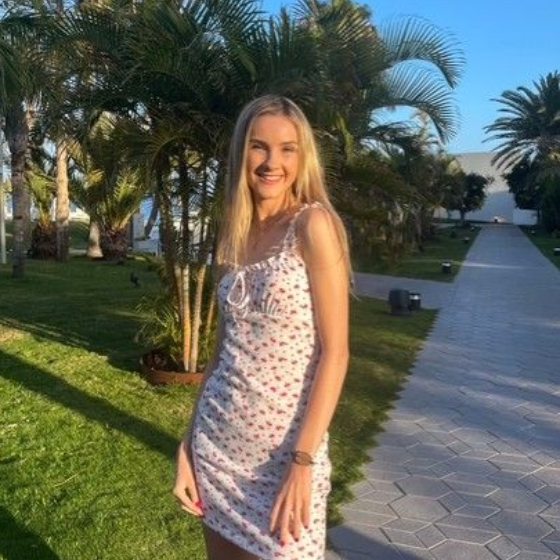 A person wearing a navy tank top and floral cardigan smiles while standing on a balcony overlooking a city and coastline.