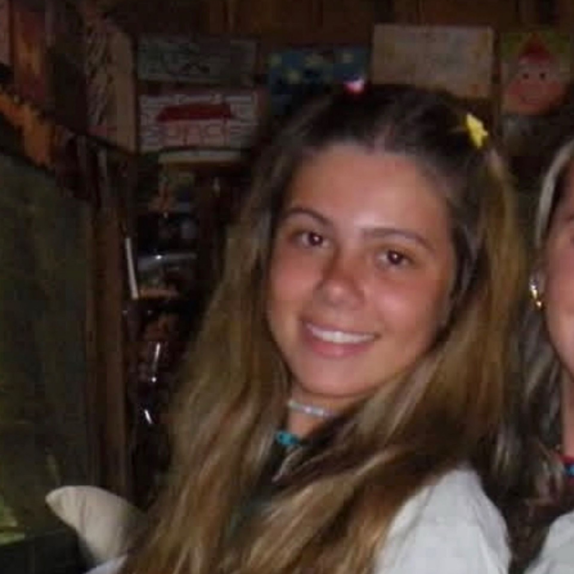 A person wearing a navy tank top and floral cardigan smiles while standing on a balcony overlooking a city and coastline.