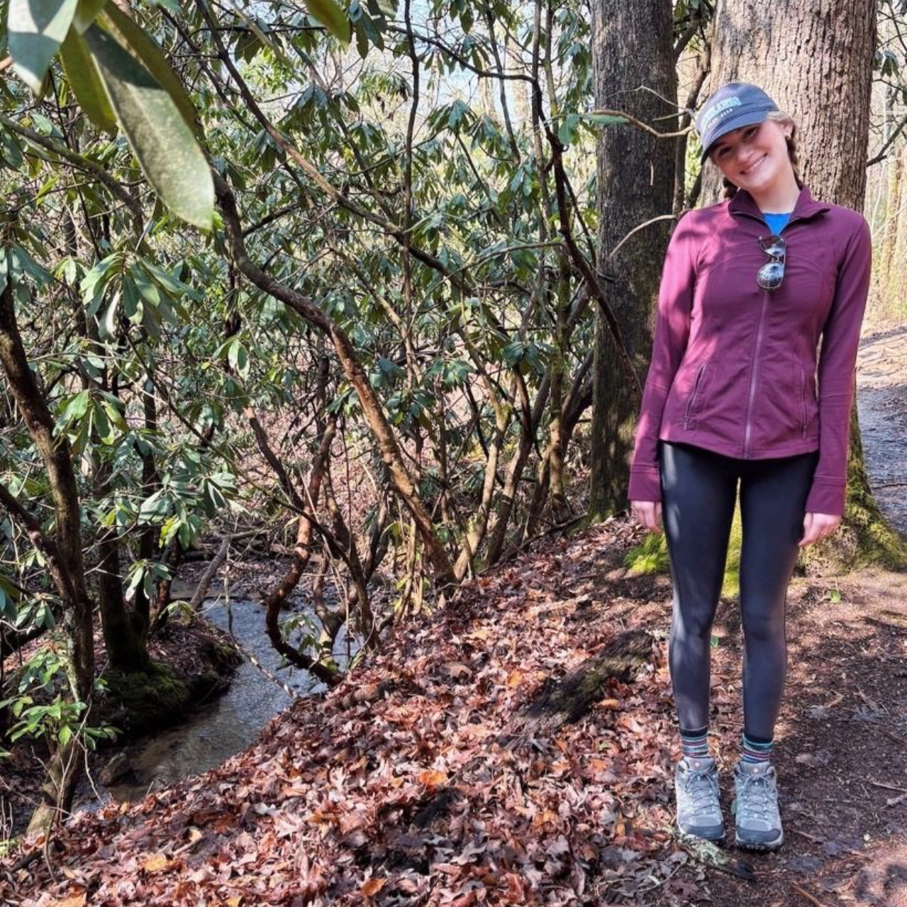 A person wearing a navy tank top and floral cardigan smiles while standing on a balcony overlooking a city and coastline.