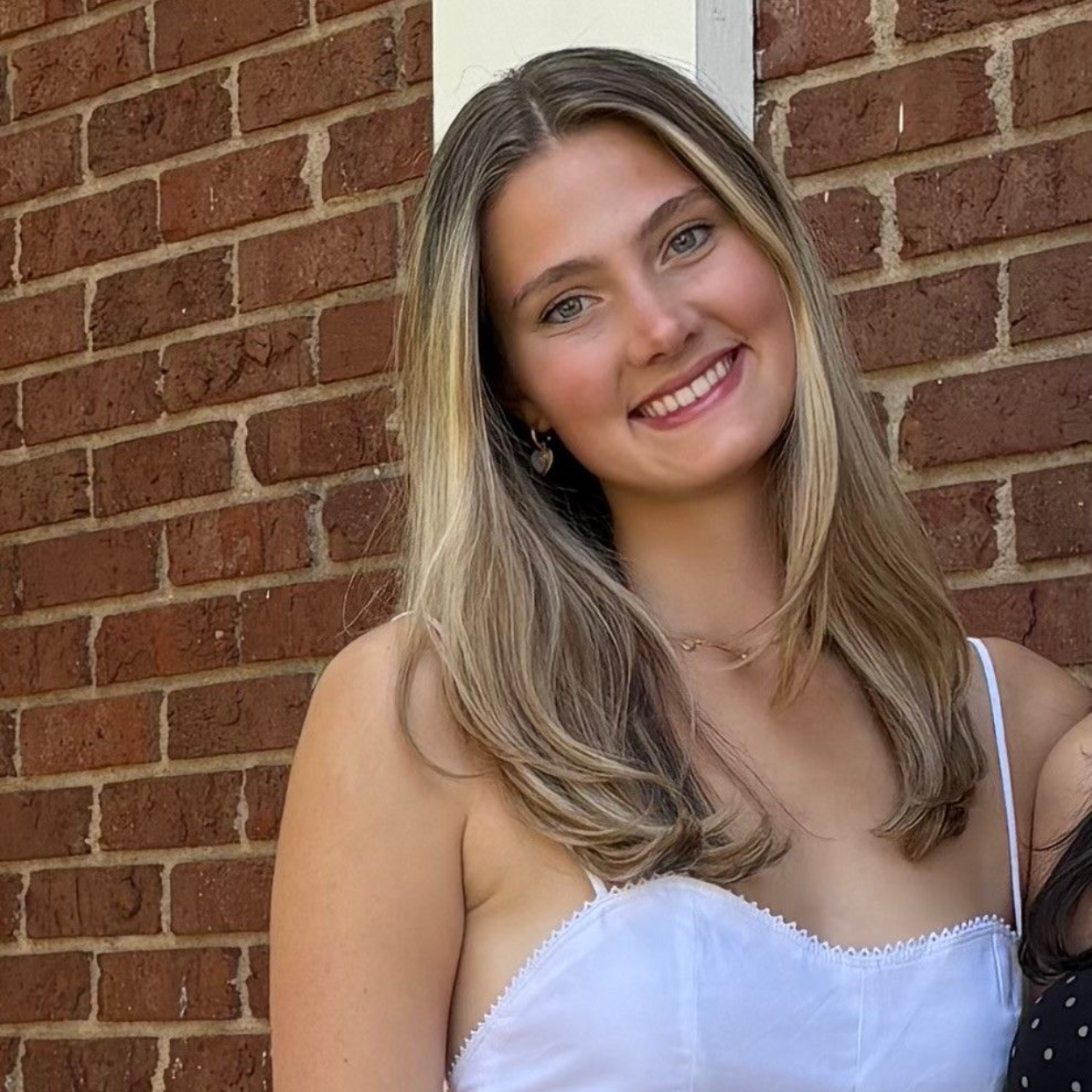 A person wearing a navy tank top and floral cardigan smiles while standing on a balcony overlooking a city and coastline.