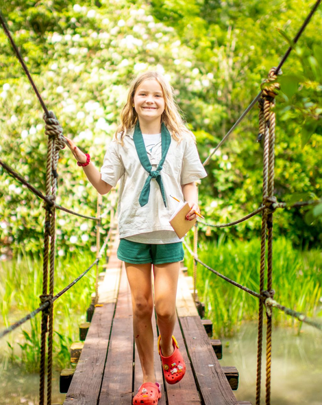 A person in a light shirt and green scarf smiles while walking across a wooden suspension bridge in a lush forest.