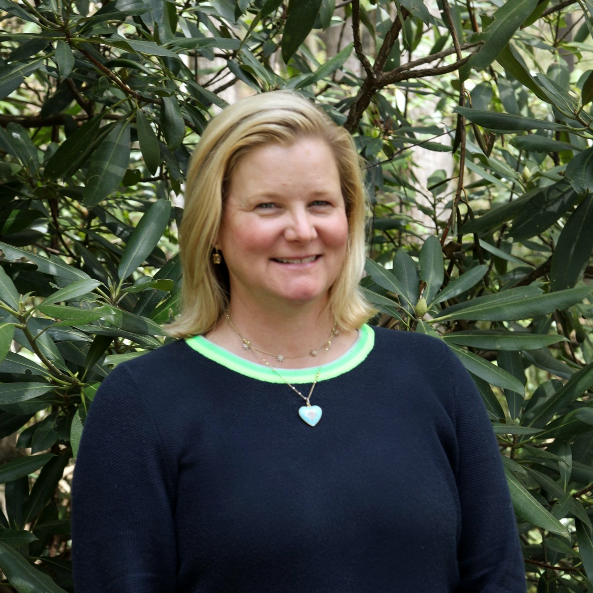 Smiling woman with blonde hair, wearing a denim jacket and white blouse, posed outdoors against a soft green background.