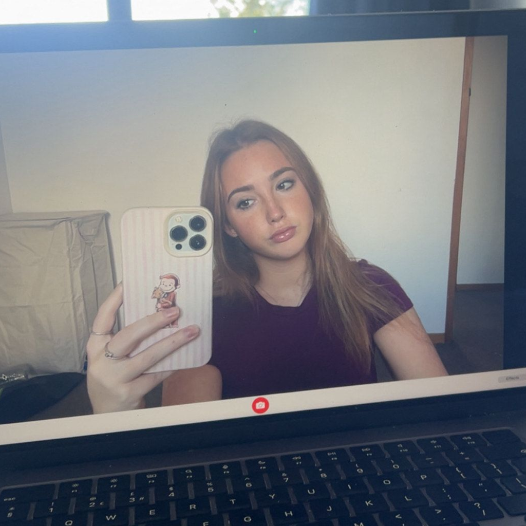 A person wearing a navy tank top and floral cardigan smiles while standing on a balcony overlooking a city and coastline.