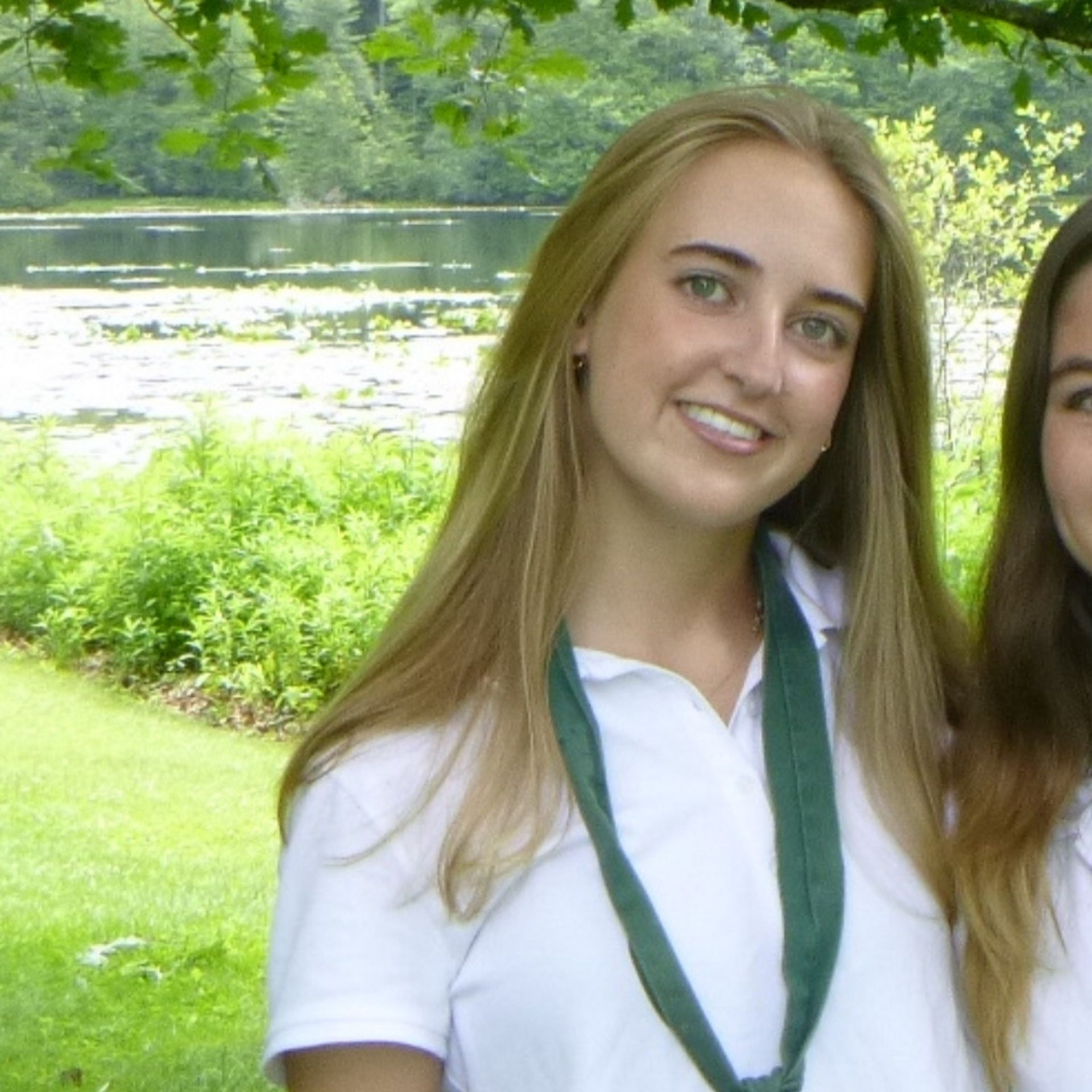 A person wearing a navy tank top and floral cardigan smiles while standing on a balcony overlooking a city and coastline.