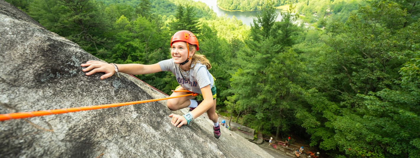 A person wearing a helmet climbs a rocky cliff face secured by an orange rope, with a lush forest and lake below.