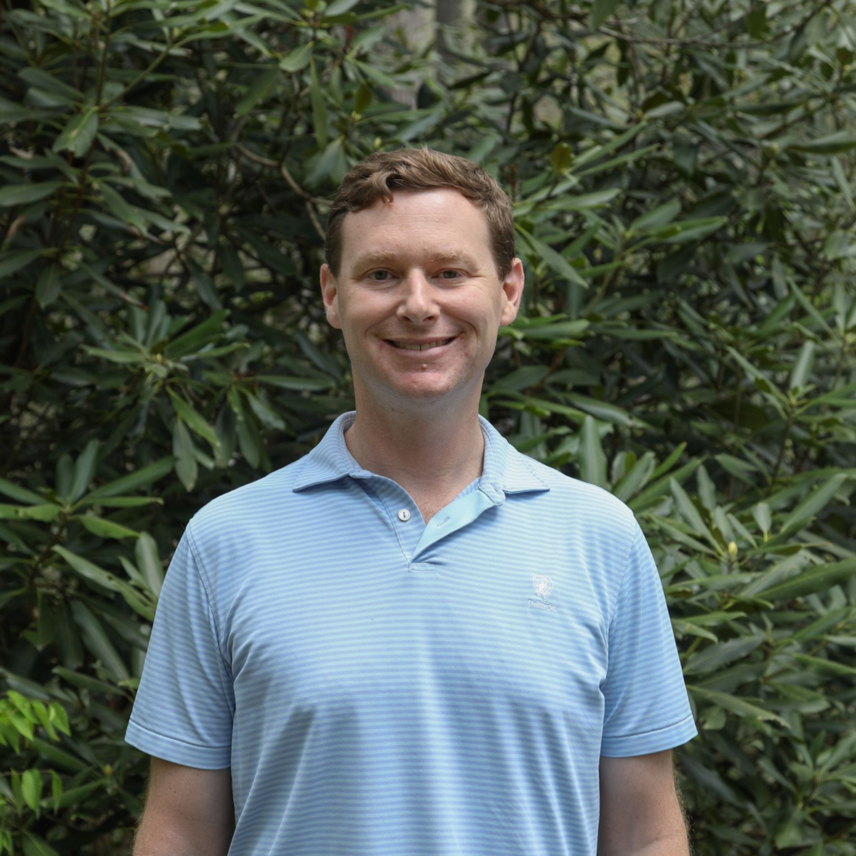 A man in a black tuxedo and bow tie smiling against a background of trees and mountains.
