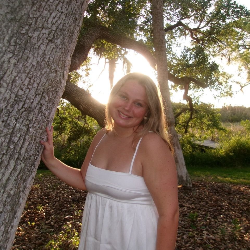 A person wearing a navy tank top and floral cardigan smiles while standing on a balcony overlooking a city and coastline.