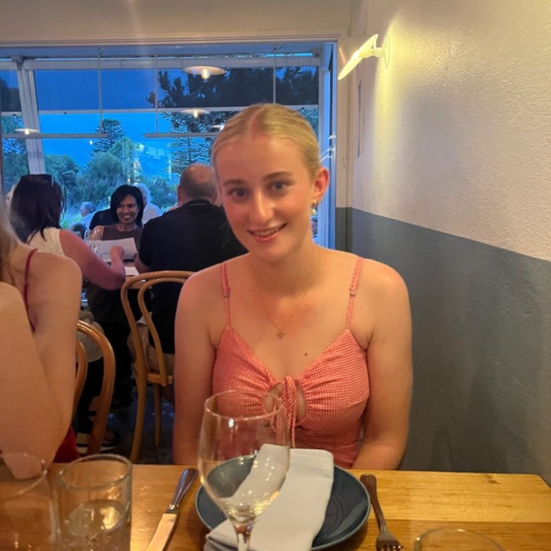 A person wearing a navy tank top and floral cardigan smiles while standing on a balcony overlooking a city and coastline.
