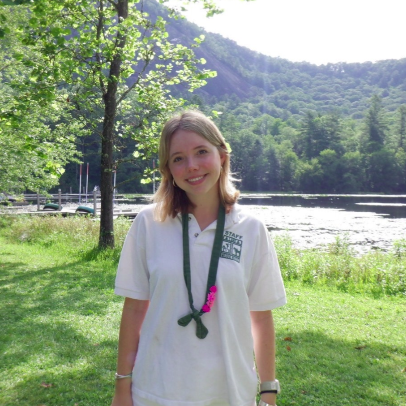 A person smiles for a photo outdoors, wearing a pink tank top and a blue whistle necklace, with others in the background.