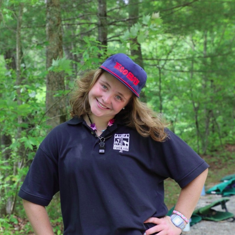 A person smiles for a photo outdoors, wearing a pink tank top and a blue whistle necklace, with others in the background.