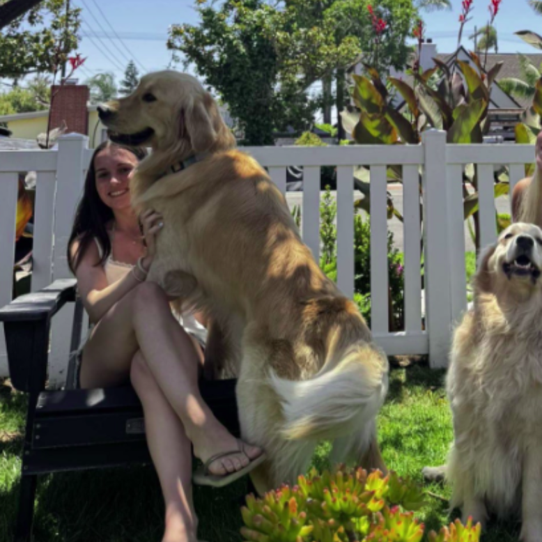 A person smiles for a photo outdoors, wearing a pink tank top and a blue whistle necklace, with others in the background.