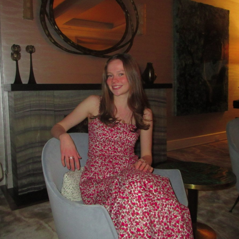 A person smiles for a photo outdoors, wearing a pink tank top and a blue whistle necklace, with others in the background.