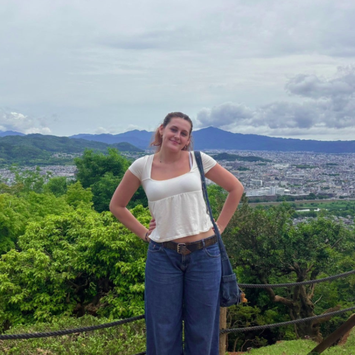 A person smiles for a photo outdoors, wearing a pink tank top and a blue whistle necklace, with others in the background.