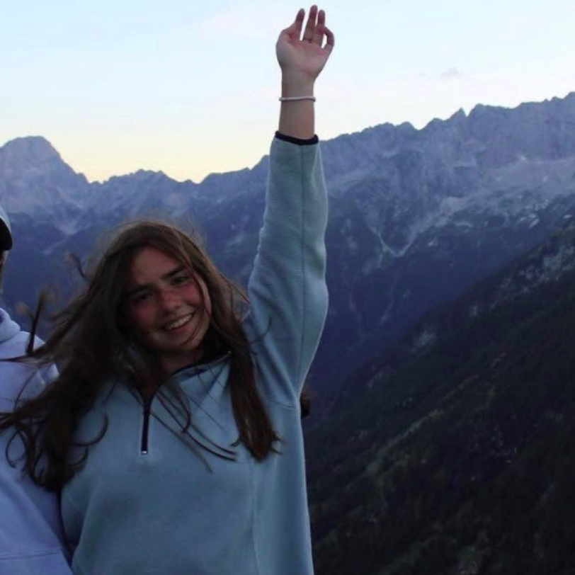 A person smiles for a photo outdoors, wearing a pink tank top and a blue whistle necklace, with others in the background.