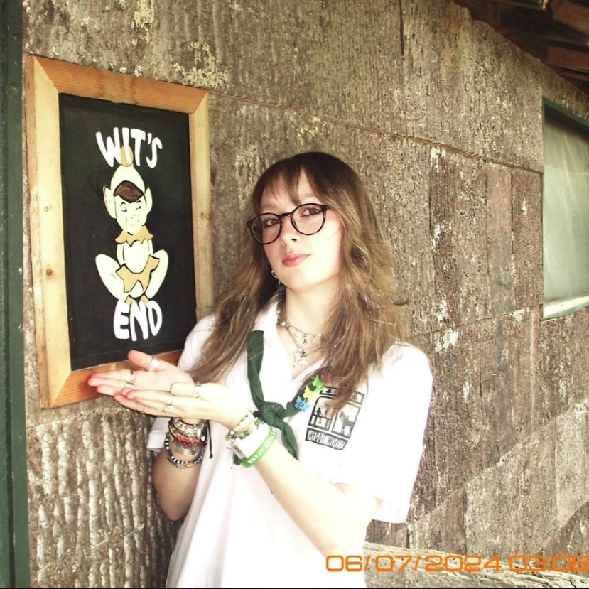 A person smiles for a photo outdoors, wearing a pink tank top and a blue whistle necklace, with others in the background.