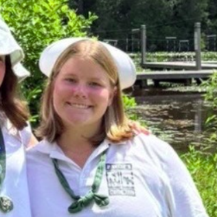 A person smiles for a photo outdoors, wearing a pink tank top and a blue whistle necklace, with others in the background.