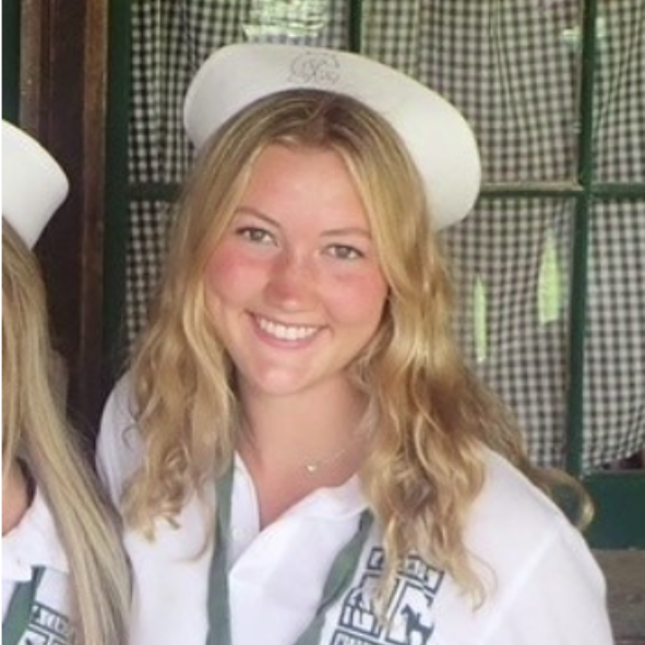 A person smiles for a photo outdoors, wearing a pink tank top and a blue whistle necklace, with others in the background.