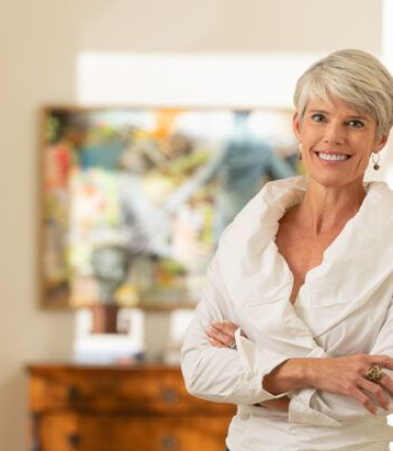 A person with short grey hair and a white top stands with arms crossed, smiling in front of a blurry abstract painting.