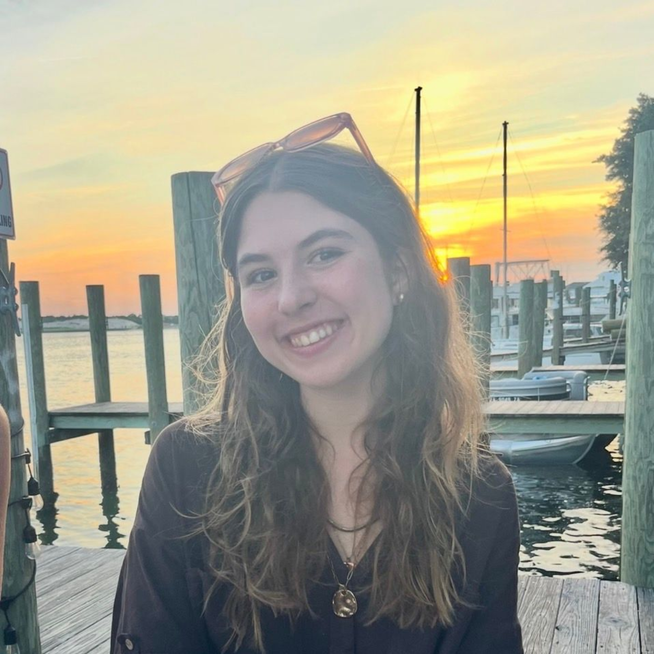 A person wearing a navy tank top and floral cardigan smiles while standing on a balcony overlooking a city and coastline.