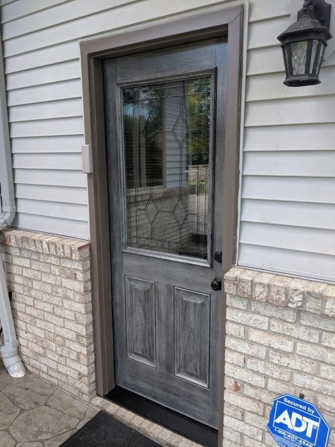 A gray door with a stained glass window is on the side of a brick house.