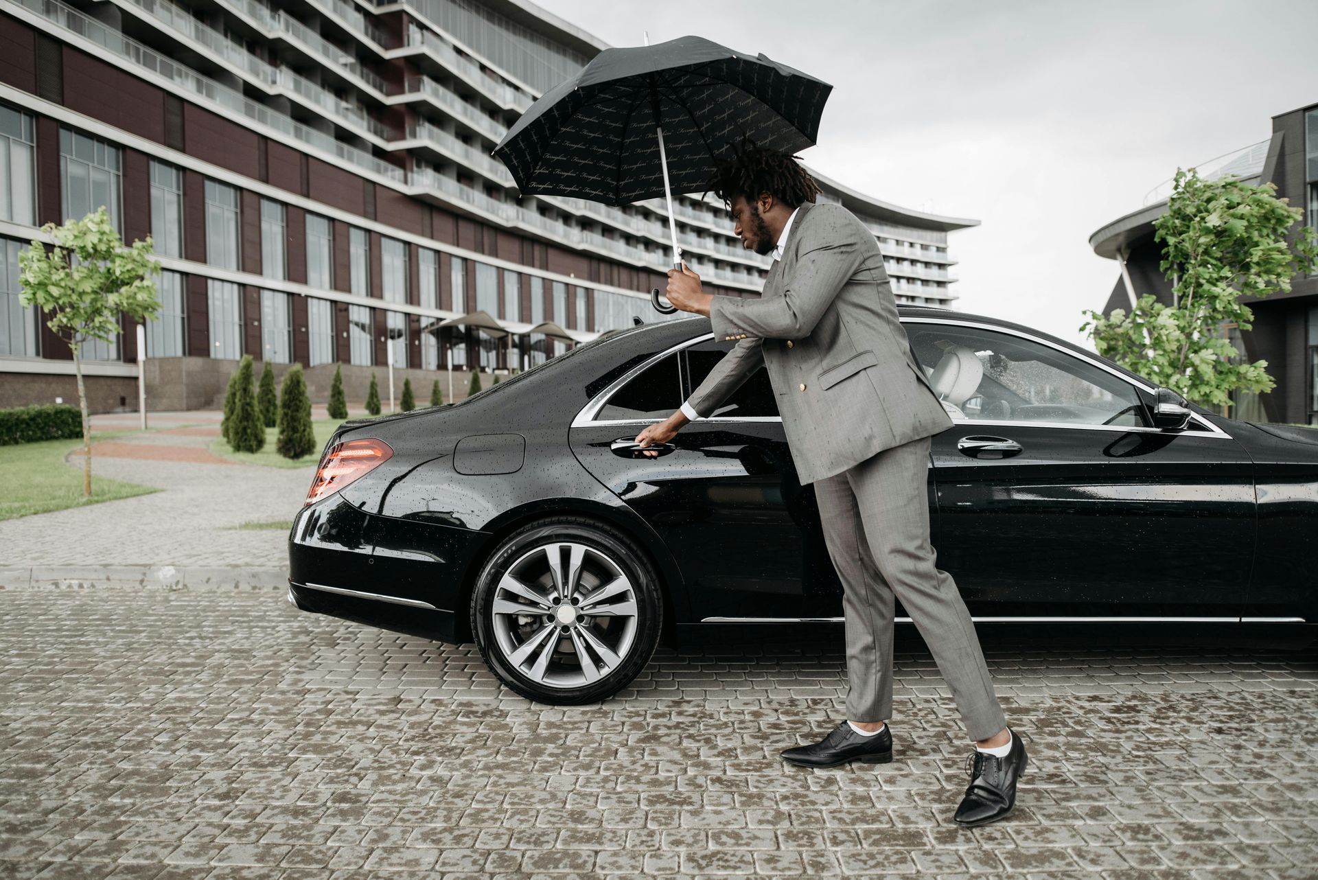 A man in a suit is opening the door of a car while holding an umbrella.