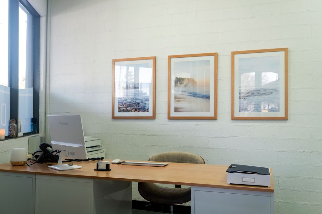 Desk With Framed Art, Computer, and Chair Against a White Brick Wall — Dr John Salmon, Kiama Family Dental In Kiama, NSW