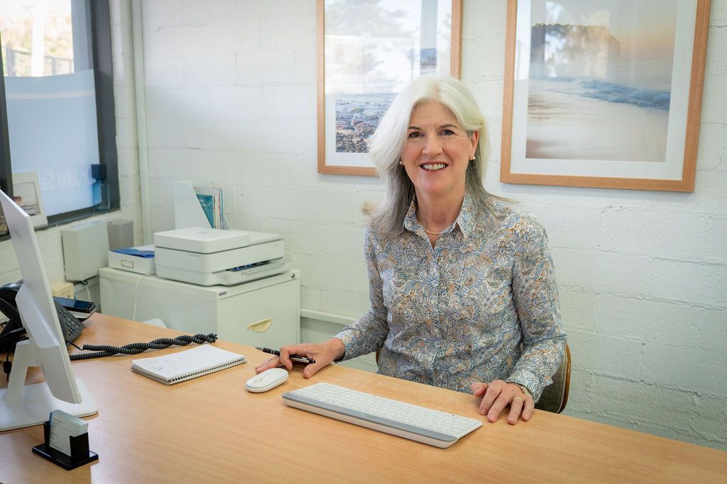 Woman With Long Grey Hair Smiles at the Camera, Sitting at a Desk — Dr John Salmon, Kiama Family Dental In Kiama, NSW